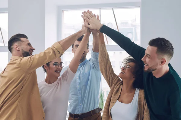 Grupo de jóvenes felices en ropa casual inteligente tomados de la mano y sonriendo mientras se reúnen en la oficina — Foto de Stock