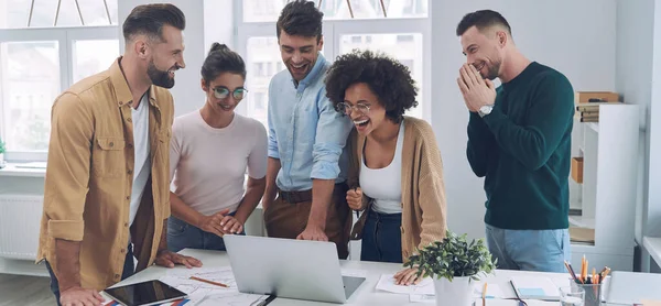 Grupo de jóvenes felices en ropa casual inteligente mirando a la computadora portátil y haciendo gestos mientras se reúnen en la oficina —  Fotos de Stock