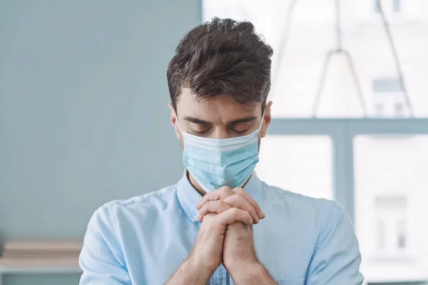 Handsome young man in protective face mask praying with his hands clasped and eyes closed — Stock Photo, Image