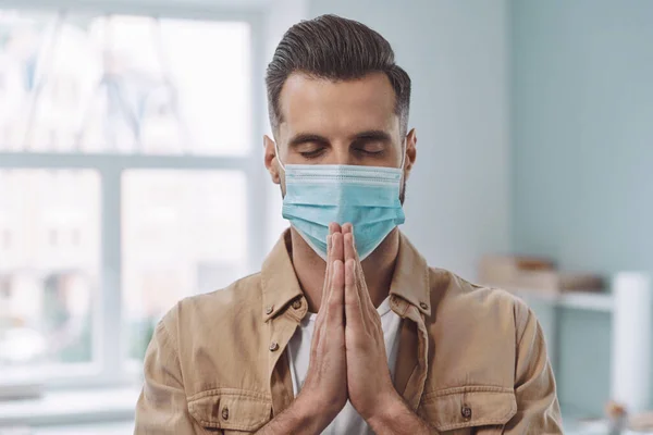 Handsome young man in protective face mask praying with his hands clasped and eyes closed — Stock Photo, Image