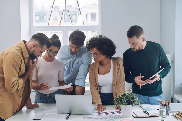 Grupo de jóvenes confiados en ropa casual inteligente discutiendo negocios mientras están de pie cerca del escritorio en la oficina —  Fotos de Stock