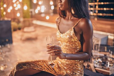 Beautiful young African woman in evening gown holding flute with champagne while spending time on night party clipart