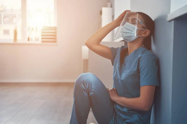 Enfermera joven cansada en ropa de trabajo protectora tocándose la cabeza mientras trabajaba en el hospital —  Fotos de Stock
