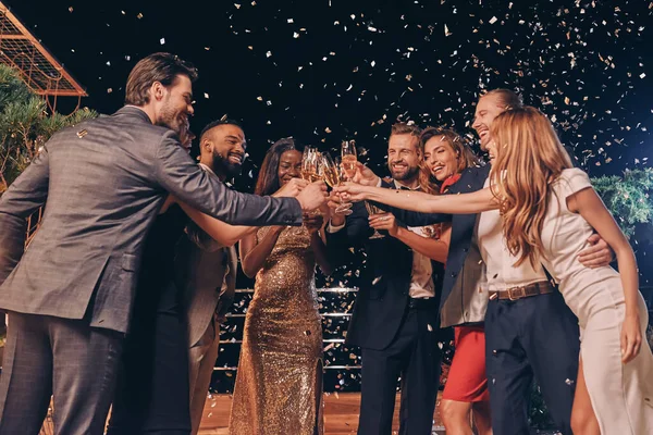 Gruppe schöner Menschen in Formalbekleidung prostet mit Champagner zu und lächelt, während Konfetti um sie herum fliegt — Stockfoto