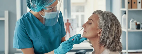 Młoda sanitariuszka używająca wymazu z nosa do wykonania testu na obecność koronawirusu u u dojrzałej kobiety podczas pracy w szpitalu — Zdjęcie stockowe