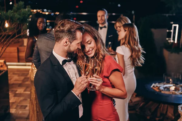 Hermosa pareja joven en ropa formal sosteniendo flautas de champán y sonriendo mientras pasa tiempo en una fiesta de lujo — Foto de Stock