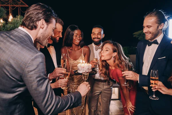 Красивая молодая женщина в вечернем платье задувает свечи на торте во время празднования дня рождения с друзьями — стоковое фото