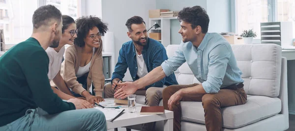 Sekelompok pemuda modern mendiskusikan bisnis dan tersenyum sambil duduk di kantor bersama — Stok Foto