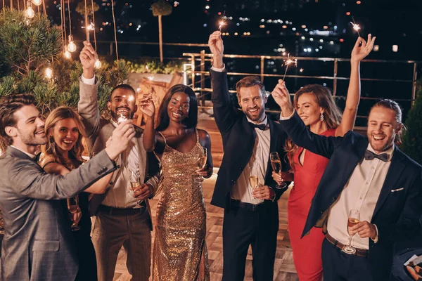 Grupo de pessoas bonitas em formalwear segurando sparklers e sorrindo enquanto passa o tempo em festa ao ar livre — Fotografia de Stock