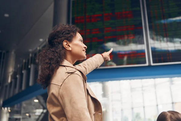 Schöne reife Frau zeigt im Flughafenterminal auf die Fluginformationstafel — Stockfoto