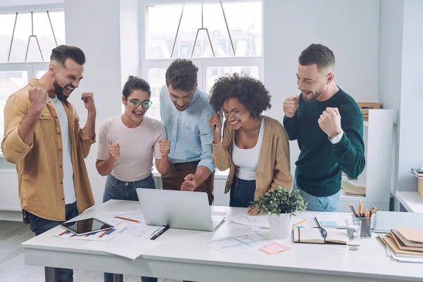 Grupp glada unga människor i smarta casual wear tittar på laptop och gester samtidigt som de träffas på kontoret — Stockfoto