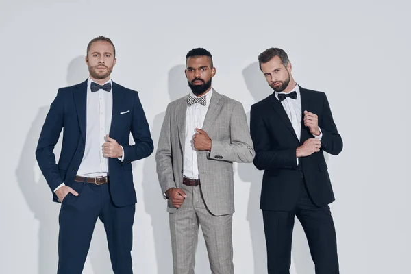 Three handsome young men in suits and bowties looking at camera while standing against gray background — Stock Photo, Image