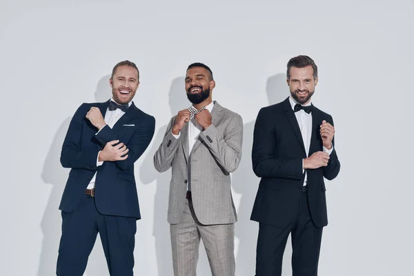 Three handsome young men in suits and bowties looking at camera and smiling while standing against gray background — Stock Photo, Image