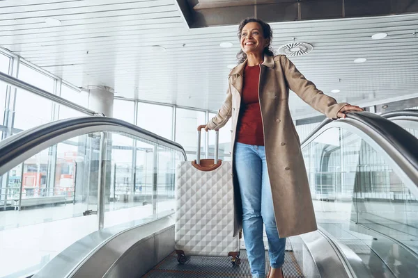 Hermosa mujer madura sonriendo y mirando hacia otro lado mientras se mueve por escaleras mecánicas en la terminal del aeropuerto —  Fotos de Stock