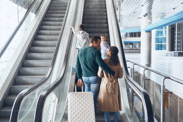 Familia feliz con dos niños pequeños moviéndose por escaleras mecánicas en la terminal del aeropuerto — Foto de Stock