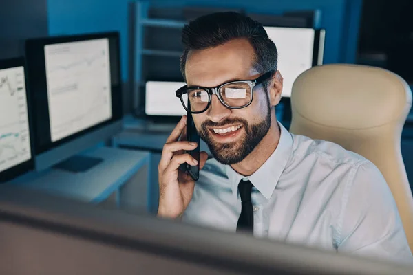 Jovem feliz falando no celular e olhando para o computador enquanto fica até tarde no escritório — Fotografia de Stock