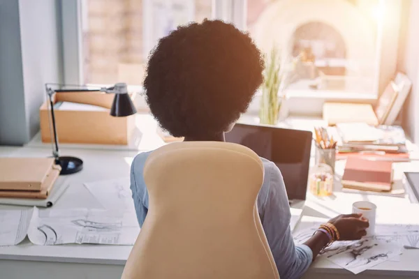 Vista posteriore della giovane donna africana che tiene la tazza con la bevanda mentre siede al suo posto di lavoro nello studio di design — Foto Stock