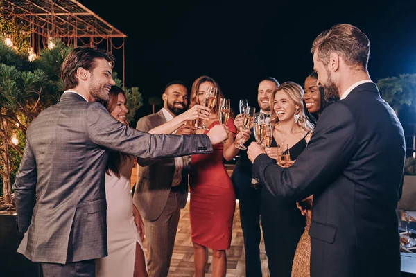 Egy csapat boldog ember ünnepi ruhában pezsgővel és mosollyal, miközben luxusbulin vannak. — Stock Fotó