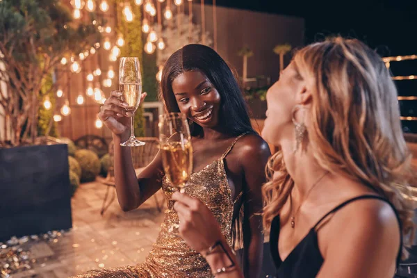 Две красивые женщины в вечерних платьях пьют шампанское и улыбаются, проводя время на роскошной вечеринке — стоковое фото