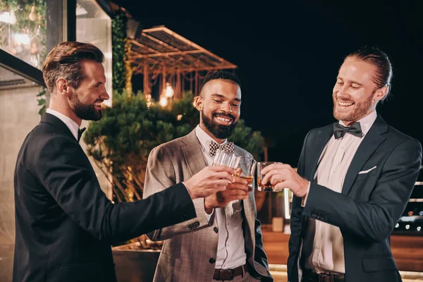 Tre bellissimi uomini in giacca e cravatta brindano con whisky e sorridono mentre trascorrono del tempo alla festa — Foto Stock