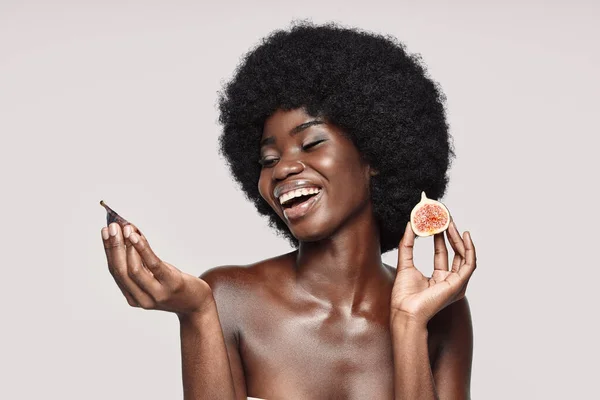 Retrato de una hermosa joven africana sosteniendo rodajas de higo y sonriendo mientras está de pie sobre un fondo gris — Foto de Stock