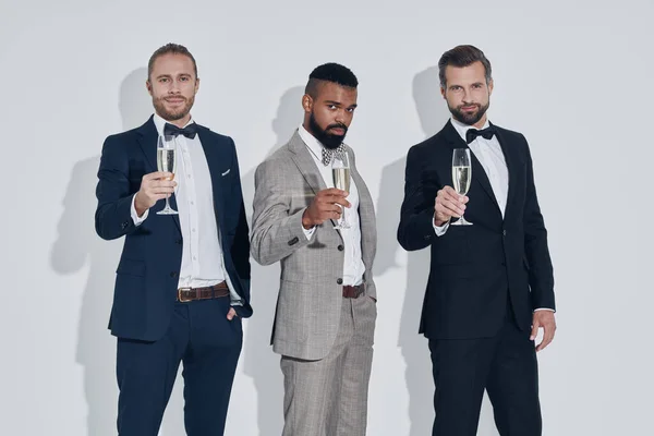 Three handsome young men in suits and bowties toasting with champagne while standing against gray background — Stock Photo, Image