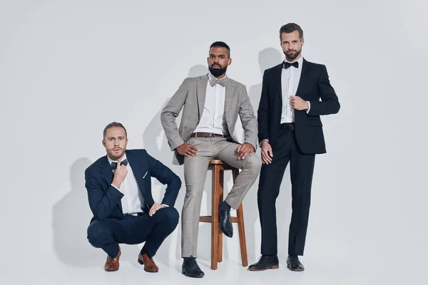 Three handsome young men in suits and bowties looking at camera while standing against gray background — Stock Photo, Image