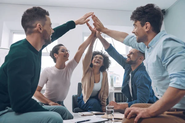 Equipo de negocios joven y seguro que mantiene las manos apretadas y sonriendo mientras se sienta en la oficina juntos — Foto de Stock