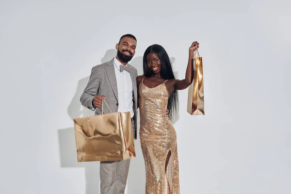 Hermosa pareja africana llevando bolsas de compras de color oro y sonriendo mientras está de pie sobre fondo gris — Foto de Stock