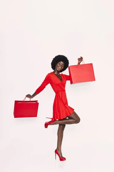 Comprimento total da bela jovem africana em vestido vermelho carregando sacos de compras vermelhos — Fotografia de Stock