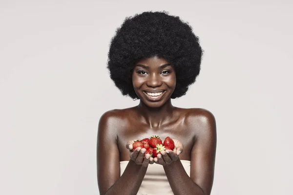 Portret van een mooie jonge Afrikaanse vrouw met aardbeien en een glimlach terwijl ze tegen een grijze achtergrond staat — Stockfoto