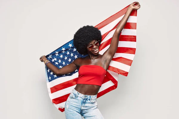 Jovem africana feliz carregando bandeira americana e sorrindo de pé contra fundo cinza — Fotografia de Stock