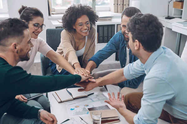 Equipe de negócios jovem confiante mantendo as mãos apertadas e sorrindo enquanto sentados no escritório juntos — Fotografia de Stock