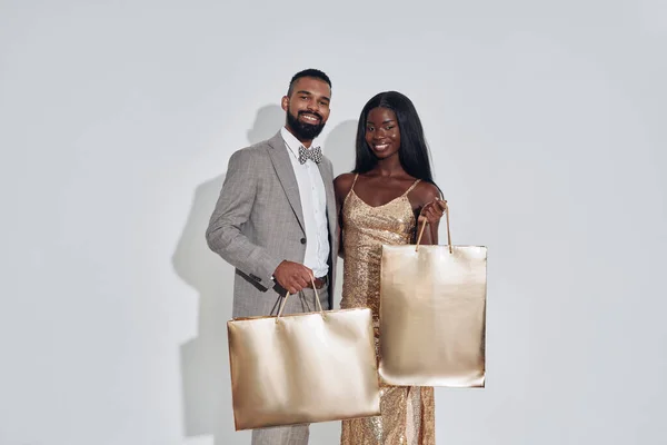 Hermosa pareja africana llevando bolsas de compras de color oro y sonriendo mientras está de pie sobre fondo gris — Foto de Stock