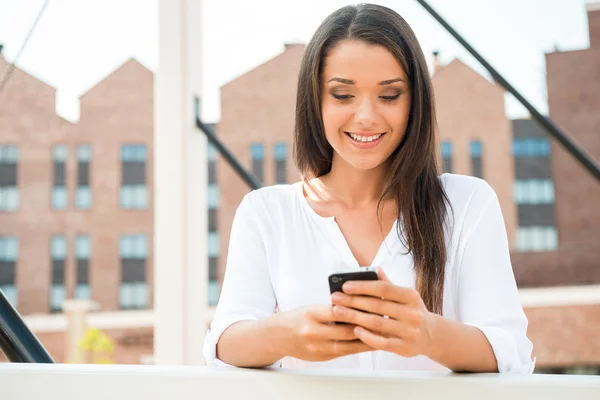 A ler a mensagem dele. Bela jovem mulher sorridente segurando telefone celular e olhando para ele enquanto está de pé ao ar livre — Fotografia de Stock