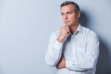 Lost in thoughts. Portrait of thoughtful mature man in shirt holding hand on chin and looking away while standing against grey background clipart