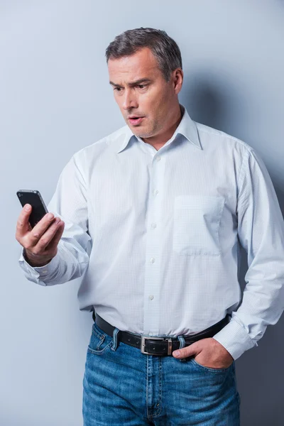 Schockierende Nachrichten. Überraschter älterer Mann im Hemd hält Handy in der Hand und schaut es sich vor grauem Hintergrund an — Stockfoto