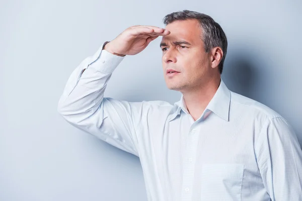 Curious mature man. Side view of curious mature man in shirt looking at view while holding hand over eyes and standing against grey background — Stock Photo, Image