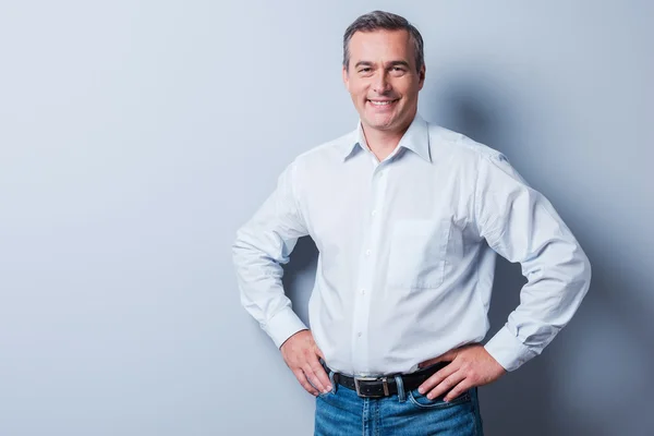 Confiado y exitoso. Hombre maduro con confianza en la camisa mirando a la cámara y sonriendo mientras se toma de la mano en la cadera y de pie sobre fondo gris —  Fotos de Stock