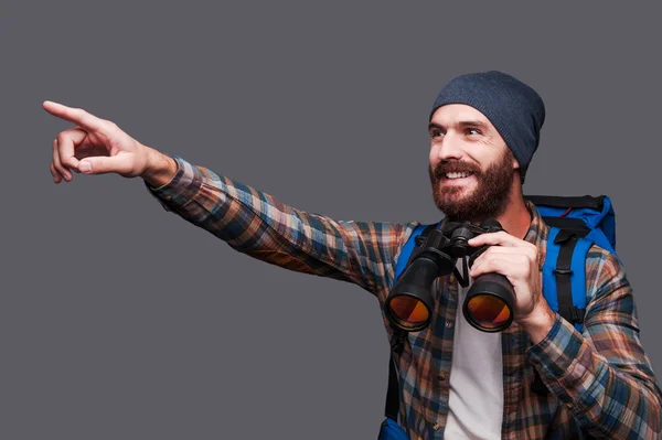 Bearded man with backpack holding binoculars and pointing away — Stock Photo, Image