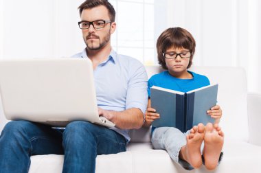 küçük oğlu kitap okuma sırasında dizüstü bilgisayar üzerinde çalışan baba