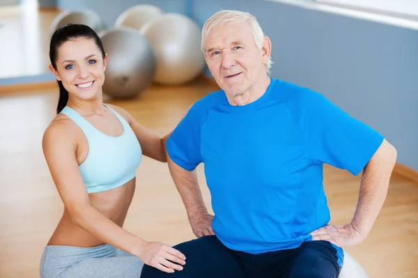 On the road to full recovery. Confident female physical therapist looking at camera and smiling while sitting close to cheerful senior man — Stock Photo, Image