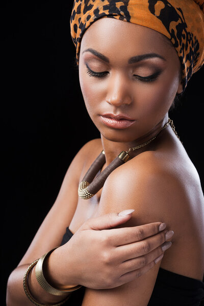 Beautiful African woman wearing a headscarf and touching her shoulder while standing against black background