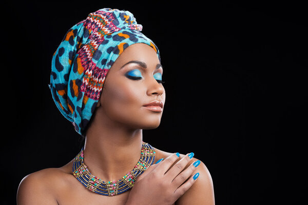 Beautiful African woman wearing a headscarf and necklace keeping eyes closed while standing against black background