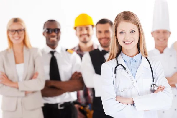 Cuando crezca seré médico. Hermosa joven doctora manteniendo los brazos cruzados y sonriendo mientras grupo de personas en diferentes profesiones de pie en el fondo — Foto de Stock