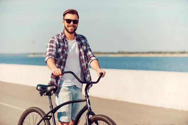 Só eu e a minha bicicleta. Bonito jovem que está perto de sua bicicleta e sorrindo enquanto está ao ar livre — Fotografia de Stock