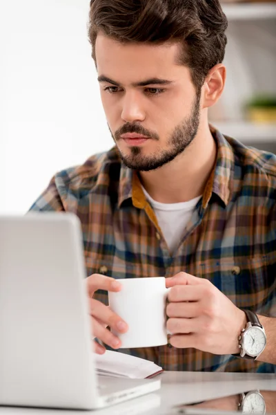 Мужчина смотрит на ноутбук и держит чашку — стоковое фото