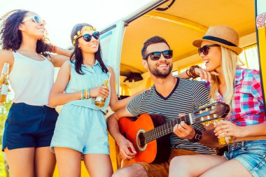 man sitting at minivan and playing guitar with three girls