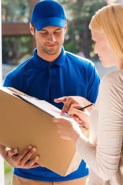 Just sign here. Handsome young delivery man holding a cardboard box while beautiful young woman putting signature in clipboard — Stockfoto