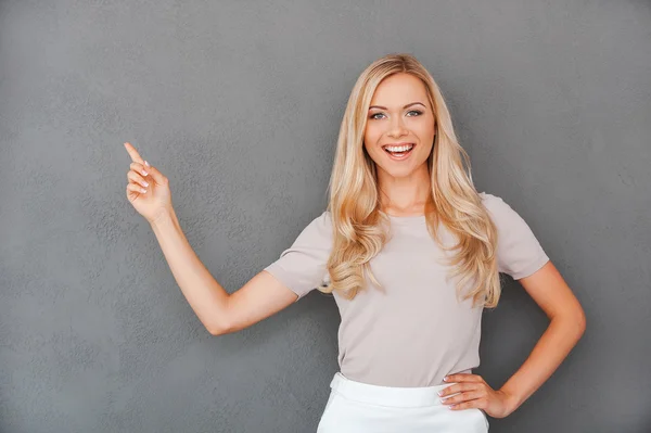 Blond hair woman pointing away and looking at camera while standing against grey background — Stok fotoğraf